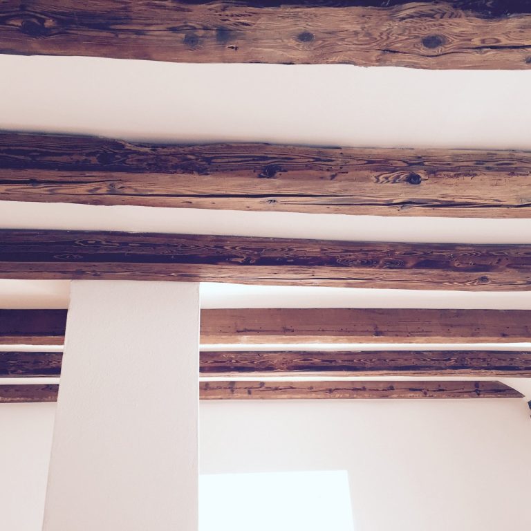 Holzdeckenbalken in einem Raum mit heller Wand und Fensterlicht.
